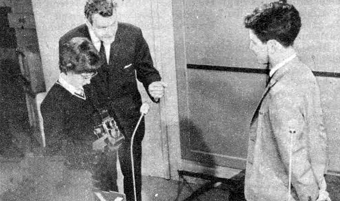 A young woman holds a camera supervised by a man; a young man looks on